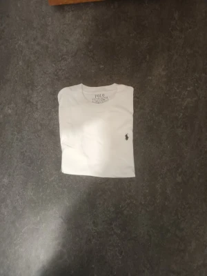 Vit t-shirt Polo Ralph Lauren - Klassisk vit t-shirt Polo Ralph Lauren med den ikoniska lilla broderade loggan på bröstet. T-shirten har rund hals och korta ärmar, tillverkad i mjuk bomull för en skön och enkel stil.