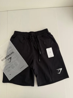 Svarta shorts från Gymshark - Svarta shorts från Gymshark med elastisk midja och snörning. Shortsen har dragkedjeförsedda fickor och en vit Gymshark-logga på ena benet. Perfekta för träning och har en sportig look.