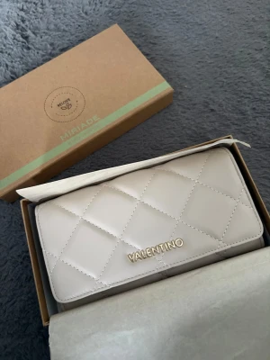 Beige quiltad plånbok Valentino - Stilren beige plånbok från Mario Valentino med quiltat mönster och guldfärgad logga framtill. Plånboken har flera fack för kort, sedlar och mynt samt stängs med tryckknapp. Tillverkad i slätt konstläder för en lyxig känsla.