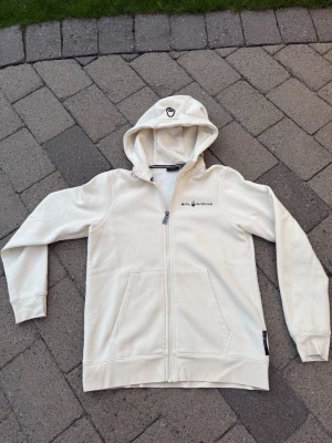 Vit hoodie från Sail Racing - Vit hoodie från Sail Racing med dragkedja framtill och två fickor. Hoodien har en broderad logga på bröstet och en liten detalj på huvan. Perfekt för en avslappnad och stilren look.