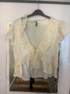 Vit spetsblus från H&M - Romantisk vit blus från H&M med vackra spetsdetaljer längs kanterna och volang vid ärmarna. Djup V-ringning och knytning framtill ger en feminin look. Tillverkad i ett lätt, glansigt tyg som känns mjukt mot huden.