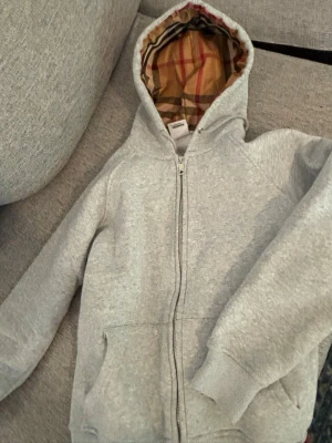 Grå zip hoodie från Burberry - Snygg grå hoodie från Burberry med dragkedja och klassiskt rutigt foder i huvan. Mjukt material och två fickor framtill. Perfekt för dig som vill ha en stilren och exklusiv look med en twist av Burberrys ikoniska mönster.