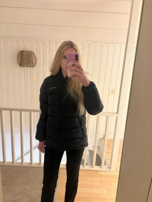 Mörkblå pufferjacka från Peak Performance - Snygg mörkblå pufferjacka från Peak Performance med huva och dragkedja framtill. Jackan har två sidofickor med dragkedja och diskret logga på bröstet. Perfekt för kalla vinterdagar tack vare det tjocka, vadderade materialet. Storleken är egentligen Small men den är lite stor i storlek upplever den som en Medium.