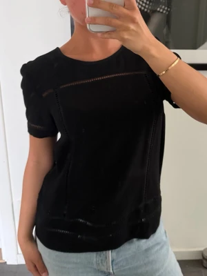 Svart topp med håldetaljer - Michael Kors strl XS - Svart topp med rund halsringning och korta ärmar. Toppen har diskreta håldetaljer längs bröstet, ärmarna och nederkanten som ger en snygg touch. Passar perfekt till jeans eller kjol för en stilren look.