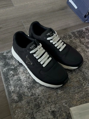 Prada practice svarta - Svarta sneakers från Prada med vit sula och vita skosnören. Skorna har en klassisk låg modell och är tillverkade i en mix av textil och läder. Sidan pryds av Pradas ikoniska metallogo. Perfekta för en stilren och trendig look.