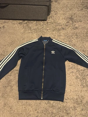 Marinblå Adidas track jacket - Snygg marinblå track jacket från Adidas med klassiska vita ränder längs ärmarna och dragkedja framtill. Jackan har ribbade muddar och två fickor på sidorna. Perfekt för en sportig och avslappnad stil.