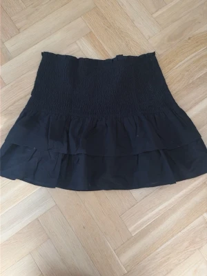 Svart volangkjol med smock - Snygg svart kortkjol med smock upptill och dubbla volanger nertill. Kjolen har en feminin och trendig look som passar perfekt till många olika outfits.