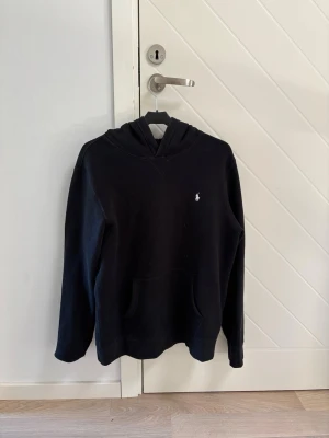 Svart hoodie från Polo Ralph Lauren - Klassisk svart hoodie från Polo Ralph Lauren med liten vit logga broderad på bröstet. Storlek 176. Mjuk och varm, perfekt för kyligare dagar.