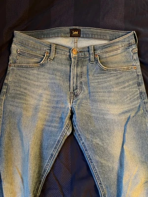 Ljusblå Lee jeans med smal passform - Snygga ljusblå jeans från Lee med klassisk femficksmodell och smal passform. Jeansen har slitningar och tvättade detaljer för en avslappnad look. Tillverkade i mjukt denimtyg med Lee-logga på bakfickan och läderpatch i midjan.