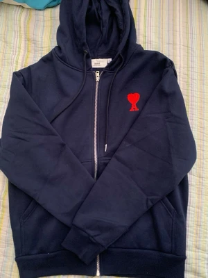 Mörkblå zip hoodie från Ami Paris - Snygg mörkblå hoodie från Ami Paris med dragkedja och huva. På bröstet sitter Ami:s ikoniska röda hjärta med A-broderi. Hoodien har fickor framtill och justerbara snören i huvan. Perfekt för en chill och stilren look.