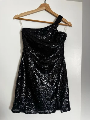 Svart glittrig oneshoulder klänning - Säljer en svart kortklänning med glittriga paljetter och oneshoulder-design. Liten slits framtill. Använd 1 gång! Från Nelly x Glamorous.