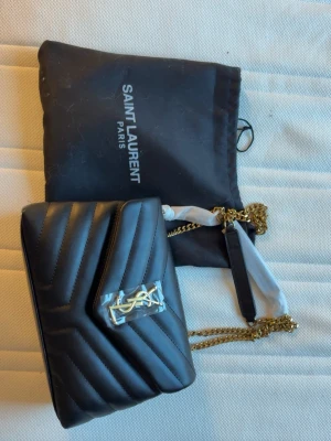Svart Saint Laurent handväska - Snygg svart handväska från Saint Laurent i quiltat läder med guldfärgad kedjerem och YSL-logga framtill. Väskan har kuvertformad lockstängning med magnetknapp och kommer med original dustbag. Perfekt för dig som vill ha en lyxig accessoar.