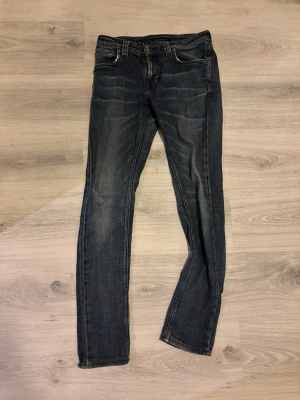 Mörkblå slim fit jeans från Nudie - Säljer ett par mörkblå slim fit jeans från Nudie med klassiska fem fickor och snygga slitningar framtill. Jeansen har kontrastsömmar, läderpatch bak i midjan och raka ben. Perfekta för en avslappnad och trendig look.  Säljer då de inte passar mig längre, storlek 29/32, kan tänka att sänka till 200