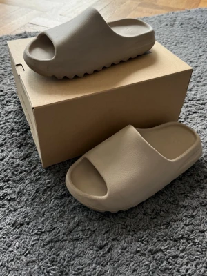 Beige Yeezy Slide tofflor - Säljer ett par Yeezy Slide tofflor i beige färg. De har en chunky, vågformad sula och är tillverkade i ett mjukt, skummaterial för maximal komfort. Perfekta för en avslappnad och trendig look.