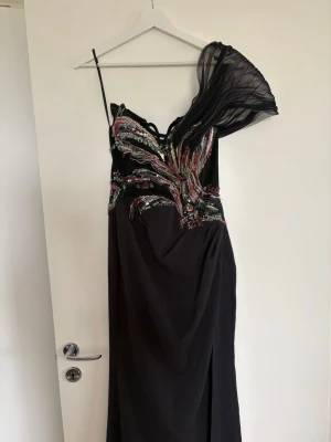Svart långklänning med paljettdetaljer - Elegant svart långklänning med en axel och transparent volangärm. Klänningen har färgglada paljettdetaljer över bysten och midjan samt en hög slits framtill. Perfekt för dig som vill sticka ut på bal eller fest. Snygg passform och unik design.