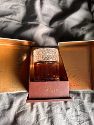Sharaf Blend Extrait de Parfum Zimaya - Lyxig parfym från Zimaya, Sharaf Blend, i en elegant glasflaska med mönstrat lock. Flaskan har en djup bärnstensfärgad vätska och levereras i en exklusiv presentförpackning med gulddetaljer. Perfekt för dig som gillar exklusiva dofter och snygg design. Nästan oanvänd och har säkert 95-98 ML kvar