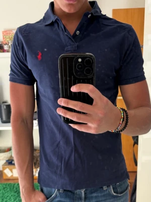 Marinblå pikétröja från Polo Ralph Lauren - Klassisk marinblå pikétröja från Polo Ralph Lauren med korta ärmar och röd broderad logga på bröstet. Tröjan har krage, knappar vid halsen och är tillverkad i mjuk bomull. Perfekt för en stilren och avslappnad look.