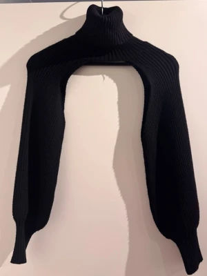 Svart stickad bolero med polokrage - Cool svart stickad bolero med hög polokrage och långa ärmar. Materialet är ribbstickat och ger en snygg struktur. Perfekt att slänga över en t-shirt eller klänning för extra edge. Märke ej angivet.
