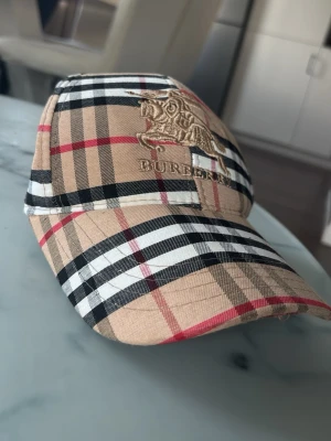 Burberry keps med klassiskt rutmönster - Snygg keps från Burberry med det ikoniska beigea, svarta, vita och röda rutmönstret. Framtill finns en broderad logga med riddare och texten Burberry. Kepsen har böjd skärm och justerbar passform. Perfekt för dig som vill ha en stilren accessoar.