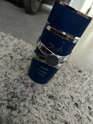 Asad Zanzibar parfym från Lattafa 100ml - Snygg parfymflaska i mörkblått med silvriga detaljer och arabiska inskriptioner. Märket är Lattafa och doften heter Asad Zanzibar. Flaskan har en unik design med metallband runt om och en dekorativ medaljong framtill.100ml