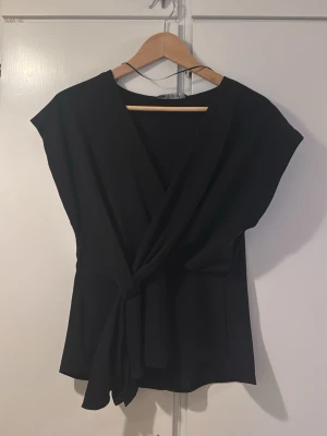 Svart omlottblus med knyt i midjan - Stilren svart omlottblus från H&M med knytband i midjan och v-ringad hals. Blusen är ärmlös och har en mjuk, lätt polyesterkvalitet som ger ett snyggt fall. Perfekt för dig som gillar minimalistisk och modern stil. Helt ny med prislapp 
