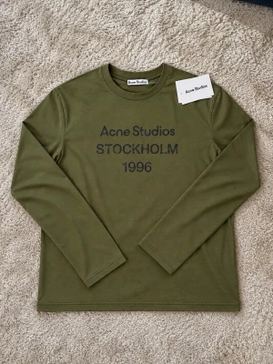 Acne Studios tröja - Snygg olivgrön långärmad tröja från Acne Studios med tryck 'STOCKHOLM 1996' på bröstet. Tröjan har rund halsringning och är gjord i mjuk bomull. Perfekt för en clean och avslappnad stil. Skulle säga att detta lätt är en av mina favorit färger på acne tröjor. Den är S men sitter som M och använd 3 gånger. Skriv om du har frågor!