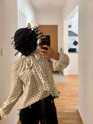 Prickig blus med rosett i halsen - Snygg vit blus med svarta prickar och stor rosett i halsen. Blusen har långärmade ärmar med lätt puff och knäppning vid ärmslut. Perfekt för en trendig och chic look. Materialet ser ut att vara ett mjukt tyg med fint fall.