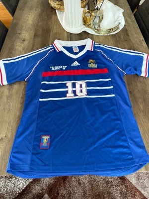 Frankrike VM 98 tröja Adidas blå - Blå fotbollströja från Adidas, officiell Frankrike VM 1998 med nummer 10 på bröstet. Tröjan har vita och röda detaljer, broderad tupp och texten 'BRESIL FRANCE 12-07-98'. Klassisk v-ringad krage med ränder. Perfekt för fotbollsfans!