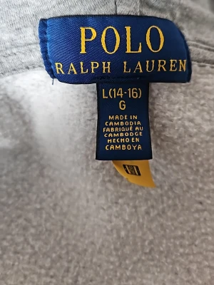 Grå hoodie från Polo Ralph Lauren - Snygg grå hoodie från Polo Ralph Lauren i storlek L (14-16). Klassisk modell med huva och mjukt material som känns skönt mot huden. Perfekt för dig som gillar enkel och stilren streetwear. Diskret logga och ribbade muddar vid ärmslut och nederkant.