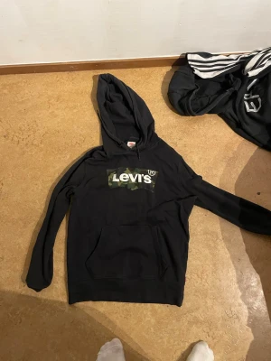 Svart Levi's hoodie med camologga - Säljer en svart hoodie från Levi's med stor kamouflagemönstrad logga på bröstet. Hoodien har huva och en klassisk magficka. Perfekt för dig som gillar streetwear och vill ha en stilren men ändå unik look. Materialet känns mjukt och skönt mot huden.