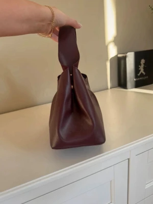 Vinröd handväska i skinn - Snygg vinröd handväska i mjukt skinn med unik form och breda handtag. Väskan har en minimalistisk design utan synliga detaljer och passar perfekt för dig som gillar stilrena accessoarer. Perfekt storlek för det viktigaste och enkel att bära i handen.