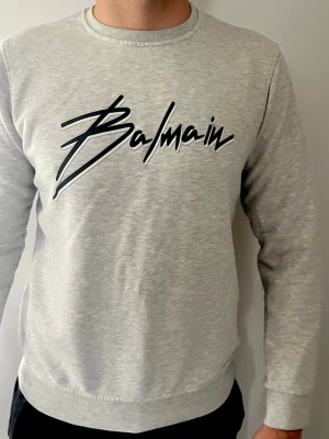 Grå sweatshirt från Balmain - Snygg ljusgrå sweatshirt från Balmain med stort svart logotryck över bröstet. Tröjan har rund halsringning och ribbade muddar vid ärmslut och nederkant. Perfekt för en avslappnad och trendig look. Storleken är XL men passformen är ENDAST M