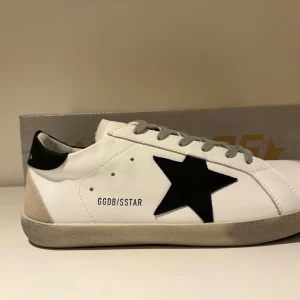 Golden Goose Superstar vita sneakers - Golden Goose Superstar sneakers i vitt läder med svart stjärndetalj på sidan, gråa skosnören och svart häl. Sulan är beige med svart mönstrad undersida. Klassisk låg modell med GGDB/SSTAR-tryck på sidan. Perfekt för en trendig streetstyle-look.