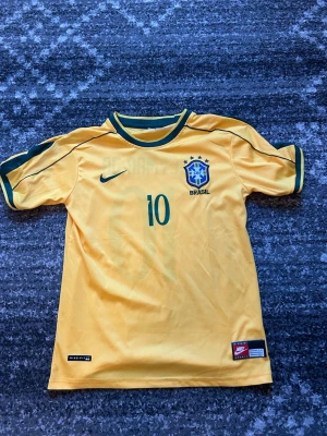 Brasilien Neymar Jr. matchtröja Nike - Gul Brasilien fotbollströja från Nike med gröna detaljer, Neymar Jr. och nummer 10 på ryggen. Officiellt klubbmärke på bröstet och tryckt Nike-logga. Tillverkad i lätt och ventilerande material, perfekt för träning eller match.