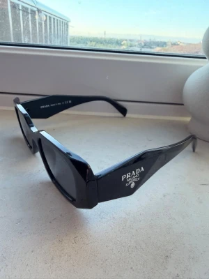 Svarta solglasögon från Prada - Snygga svarta solglasögon från Prada med breda skalmar och tydlig logga på sidan. Glasögonen har en kantig form och är tillverkade i blank plast som ger en lyxig känsla. Perfekta för dig som vill ha en statement-accessoar.