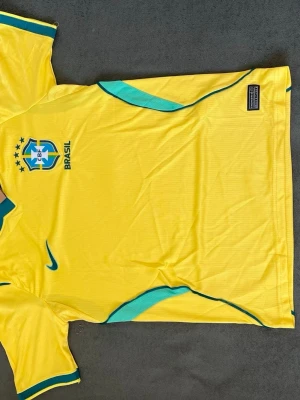 Brasilien Nike fotbollströja gul - Gul Brasilien fotbollströja från Nike med gröna detaljer och CBF-emblem på bröstet. Tröjan har rund hals med grönt kantband och Nike-logga broderad på framsidan. Tillverkad i lätt och ventilerande material, perfekt för träning eller match.