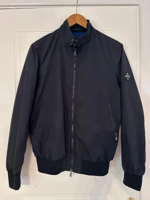 Mörkblå bomberjacka från Lyle & Scott - Snygg mörkblå bomberjacka från Lyle & Scott med hög krage och dragkedja framtill. Jackan har två snedställda fickor med dragkedja och ribbade muddar vid ärmslut och midja. Liten logotyp på ärmen ger en clean och modern look. Skriv om ni har frågor.