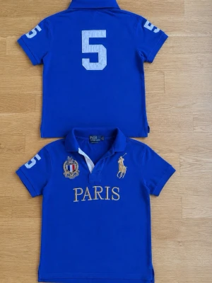 Paris  Polo Ralph Lauren - Snygg blå pikétröja från Polo Ralph Lauren med guldfärgad brodyr av loggan och texten PARIS framtill. Nummer 5 på ärmar och rygg, samt detaljerad brodyr på bröstet. Klassisk krage och korta ärmar, perfekt för en sportig look.