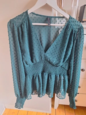 Grön prickig blus från new yorker - Trendig grön blus från new yorkee med v-ringning och transparenta långärmar. Blusen har ett prickigt strukturmönster, smock i midjan och vid ärmslut samt volang nedtill. Tillverkad i ett lätt och luftigt material som ger en snygg siluett.