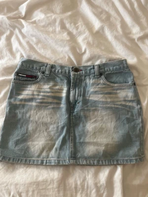 Ljusblå jeanskjol från Tommy Hilfiger - Säljer en ljusblå jeanskjol från Tommy Hilfiger med klassisk femficksdesign och slitna detaljer framtill. Kjolen har bälteshällor och knappstängning i midjan. Perfekt till sommaren för en avslappnad och trendig look. Midjemått rakt över: 37,5cm. Längd på kjolen: 36cm