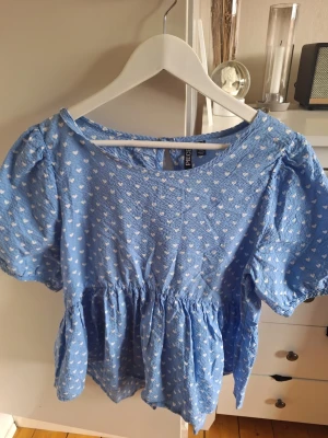 Blå blus med hjärtmönster från PIECES - Söt blå blus från PIECES med vita hjärtan över hela tyget. Blusen har puffiga korta ärmar, rund halsringning och en lätt rynkad midja som ger en avslappnad siluett. Tillverkad i ett tunt, strukturerat tyg som känns luftigt och bekvämt.