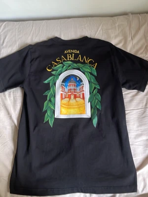 Svart Casablanca t-shirt med tryck - Snygg svart t-shirt från Casablanca med färgglatt tryck av en byggnad och gröna blad på både bröstet och ryggen. Texten 'Avenida Casablanca' syns ovanför motivet. Klassisk passform och rund hals. Perfekt för dig som gillar unika prints. Mammas död den e äkta💯