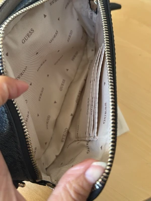 Svart handväska från Guess - Snygg svart handväska från Guess med logomönster och silverfärgad dragkedja. Invändigt finns flera fack, inklusive kortfack och innerficka med dragkedja. Väskan är fodrad med beige tyg med Guess-logga. Perfekt för att hålla ordning på dina grejer.väskan är aldrig använt.