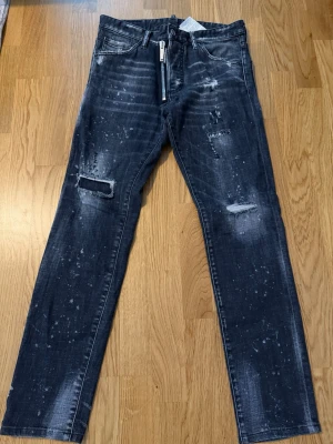 Svarta distressed jeans från Dsquared2 - Säljer ett par svarta slim fit jeans från Dsquared2 med slitningar, hål på knäna och coola färgstänk. Jeansen har klassisk femficksmodell, dragkedjegylf och är tillverkade i bomull med lite stretch. Perfekta för en edgy streetstyle-look.