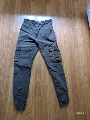 Grå cargobyxor från Madlady - Snygga grå cargobyxor från Madlady med flera praktiska fickor på benen och resår vid bensluten. Byxorna har klassisk cargomodell med knapp och dragkedja framtill samt bälteshällor. Perfekta för en avslappnad streetstil.