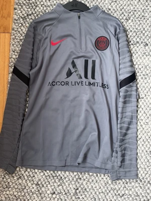 Grå PSG träningströja från Nike - Snygg grå långärmad träningströja från PSG med svarta och röda detaljer. Tröjan har halv dragkedja, PSG-logga och Nike swoosh på bröstet samt texten 'Accor Live Limitless' och 'Visit Rwanda' på ryggen. Tillverkad i funktionsmaterial som passar perfekt till träning.