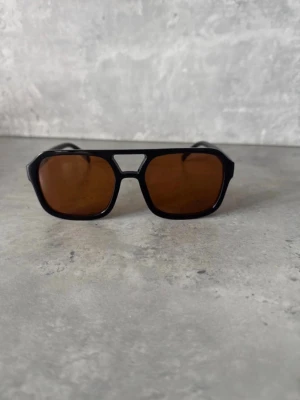 Svarta retro solglasögon med bruna glas - Snygga solglasögon med svart, kantig båge och bruna glas. Modellen har en cool retrovibe med bred brygga och markerade linjer. Perfekta för dig som vill sticka ut med en unik accessoar till sommaren. Helt nya