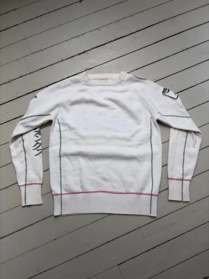 Jack & Jones stickad tröja - Vit stickad tröja med gröna och röda sömmar, samt broderade detaljer på ärmar och axel. Tröjan har rund halsringning och ribbade muddar. Unika tryck och brodyr ger en cool och personlig stil. Storlek S men passar också XS.