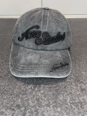 Grå keps från Acne Studios - Säljer en grå keps från Acne Studios med broderad svart logga framtill och på skärmen. Kepsen har en klassisk böjd skärm och justerbar rem bak. Tillverkad i tvättad bomull för en avslappnad look.