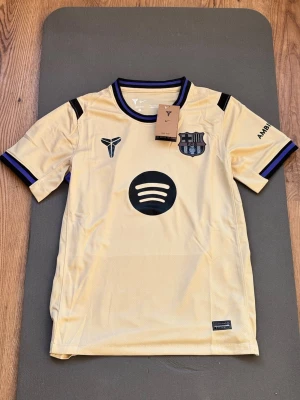 FC Barcelona - Away - Snygg gul FC Barcelona matchtröja med svarta och blå detaljer. Tröjan har klubbmärke, Nike-logga och Spotify som sponsor framtill samt UNHCR-tryck på ryggen. Tillverkad i lätt och ventilerande material, perfekt för fotboll eller träning.
